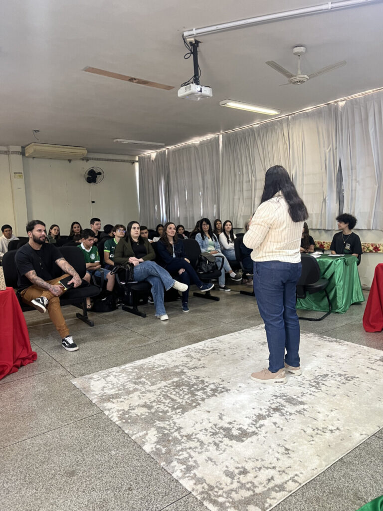 A imagem mostra uma mulher de cabelos longos e escuros, vista de costas, falando para um grupo de pessoas em uma sala de aula. Ela veste uma blusa clara listrada e calça jeans azul. À frente dela, várias pessoas estão sentadas em cadeiras, prestando atenção. O ambiente é simples, com cortinas brancas cobrindo as janelas, ventiladores de teto e um projetor suspenso. O chão é de piso frio e há um tapete claro no centro, onde a mulher está posicionada. À esquerda e à direita da imagem, mesas cobertas com tecidos vermelhos e verdes completam a decoração.