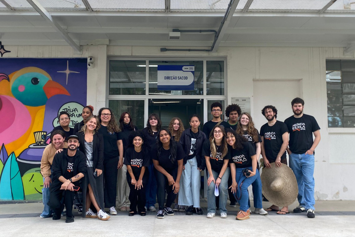 Um grupo de estudantes e integrantes da Agência Escola da UFPR posa sorrindo em frente ao prédio do SACOD, no Campus de Comunicação e Artes da Universidade Federal do Paraná. À esquerda, um mural colorido com desenhos de pássaros compõe o cenário.