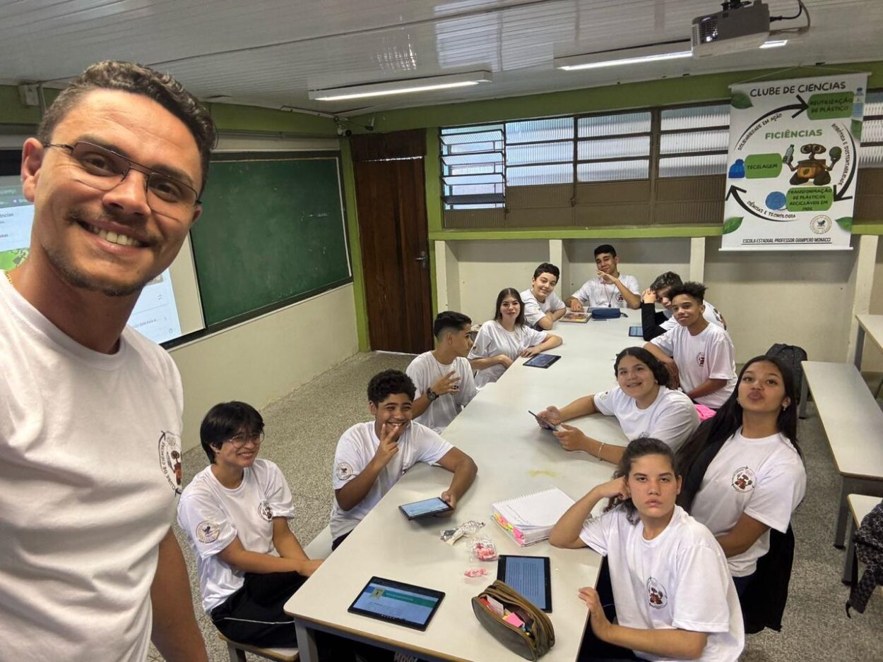 A imagem mostra uma sala de aula onde um grupo de estudantes, todos usando camisetas brancas, está reunido em torno de uma mesa retangular. Ao fundo, há um quadro verde e um cartaz na parede com o título “Clube de Ciências”, que traz ilustrações relacionadas à reciclagem e reaproveitamento de plástico. À esquerda, na frente da imagem, o professor sorri para a câmera, tirando uma selfie com os alunos. Os estudantes estão sentados em grupos, alguns segurando tablets e outros cadernos, e parecem engajados na atividade.