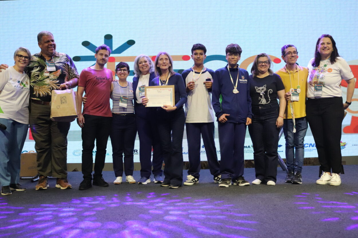 A foto mostra um grupo de pessoas reunido no palco da FECCI, entre clubistas, professores e autoridade, todos posicionadas lado a lado e olhando para a câmera. Ao centro, a professora do clube Arquitetos da Natureza segura o certificado do prêmio, enquanto os alunos do clube usam medalhas penduradas no pescoço. Algumas pessoas vestem camisetas com logos do evento ou da escola, e outras usam roupas casuais. No fundo, há um telão iluminado com elementos gráficos coloridos. A iluminação do palco deixa reflexos roxos no chão. O clima geral é de celebração e premiação.