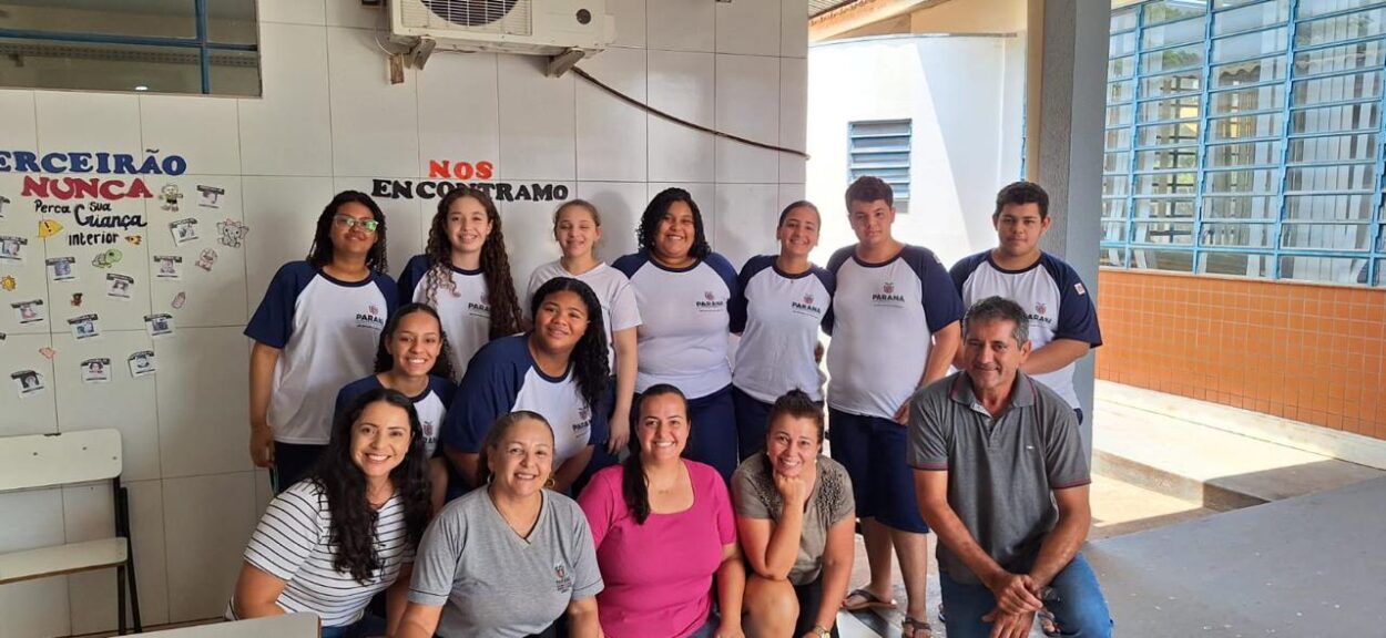 A imagem mostra um grupo de pessoas reunidas em um ambiente escolar. Na frente, há quatro mulheres e um homem agachados, enquanto ao fundo estão mais dez pessoas, entre estudantes e educadores, posando e sorrindo para a foto. Os estudantes vestem uniformes da rede estadual do Paraná, com camisetas brancas e azuis. Ao fundo, há paredes revestidas com azulejos brancos e um mural com frases e desenhos coloridos.
