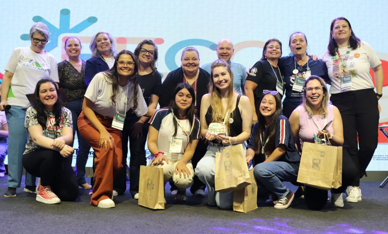 Na foto, um grupo grande de pessoas está reunido no palco da FECCi, formando duas fileiras: algumas agachadas à frente e outras em pé atrás. Todas estão sorrindo e posando para a câmera. Várias pessoas seguram sacolas de papel e crachás do evento, e a professora do clube exibe um pequeno troféu. O fundo é iluminado por um telão com elementos gráficos coloridos, criando um clima festivo. O ambiente sugere um momento de celebração e reconhecimento