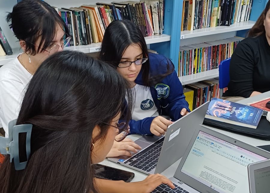 A imagem mostra as alunas do clube trabalhando, usando notebooks para escrever e estudar. Ao fundo, há estantes de livros.