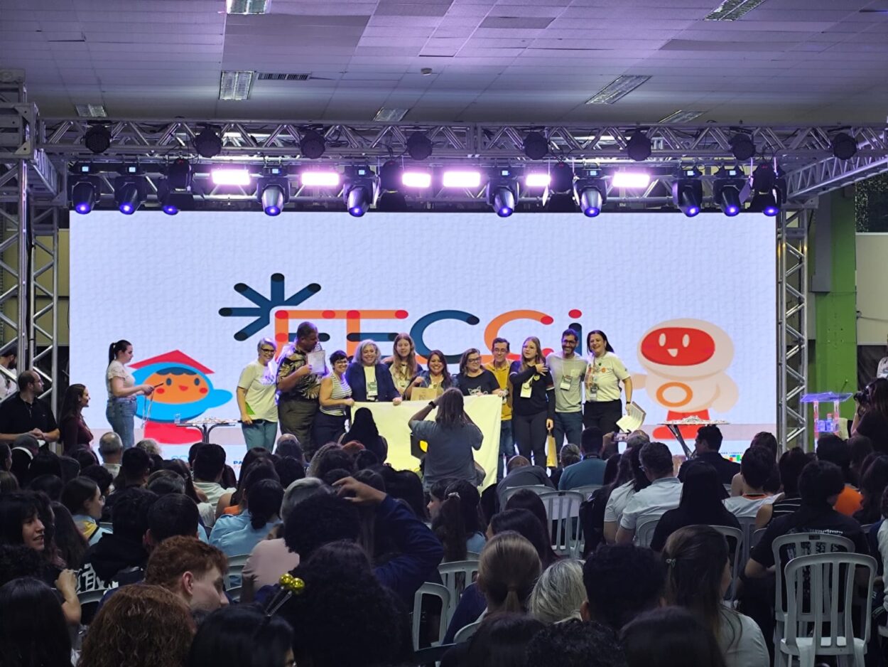 Foto mostra o palco principal da Fecci 2025, com um grande telão ao fundo exibindo o logo colorido do evento. Em frente ao telão, um grupo de professores e estudantes posam sorridentes para a foto, segurando certificados e uma faixa branca. No público, várias pessoas assistem à cerimônia sentadas em cadeiras brancas. O ambiente é iluminado por luzes de palco roxas e a plateia ocupa quase todo o espaço do salão.