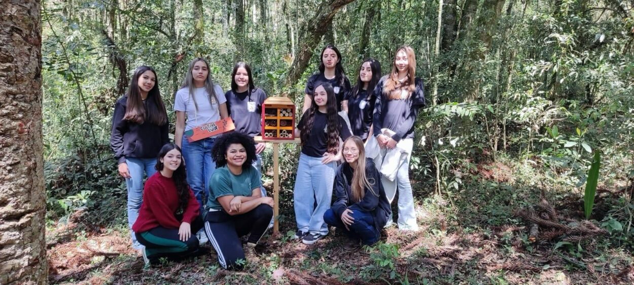 A imagem mostra um grupo de 10 estudantes em meio a uma área de floresta. Elas estão reunidas ao redor de um hotel de insetos, uma estrutura de madeira com compartimentos preenchidos por bambus, pinhas e outros materiais naturais, instalada sobre um suporte. As alunas estão em pé e agachadas, sorrindo e posando para a foto. No canto esquerdo, uma das estudantes segura uma caixa decorada. Ao fundo, é possível ver muitas árvores, vegetação densa e o chão coberto por folhas secas, reforçando o ambiente natural onde o projeto foi realizado.