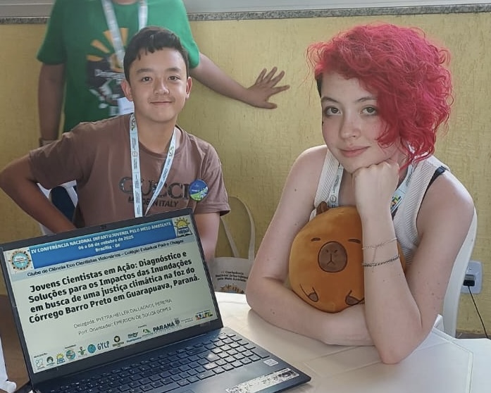 Na imagem, dois estudantes estão sentados lado a lado em uma mesa. À frente deles há um notebook aberto exibindo uma apresentação intitulada “Jovens Cientistas em Ação: Diagnóstico e Soluções para os Impactos das Inundações em busca de uma Justiça Climática na foz do Córrego Barro Preto em Guarapuava, Paraná”. A estudante à direita tem cabelo curto e vermelho, veste uma blusa clara e segura um travesseiro marrom em formato de urso. Ao lado dela, à esquerda, está um colega com camiseta marrom e crachá pendurado no pescoço, sorrindo para a câmera. Ao fundo, aparece uma parede amarela e parte de outra pessoa em pé.