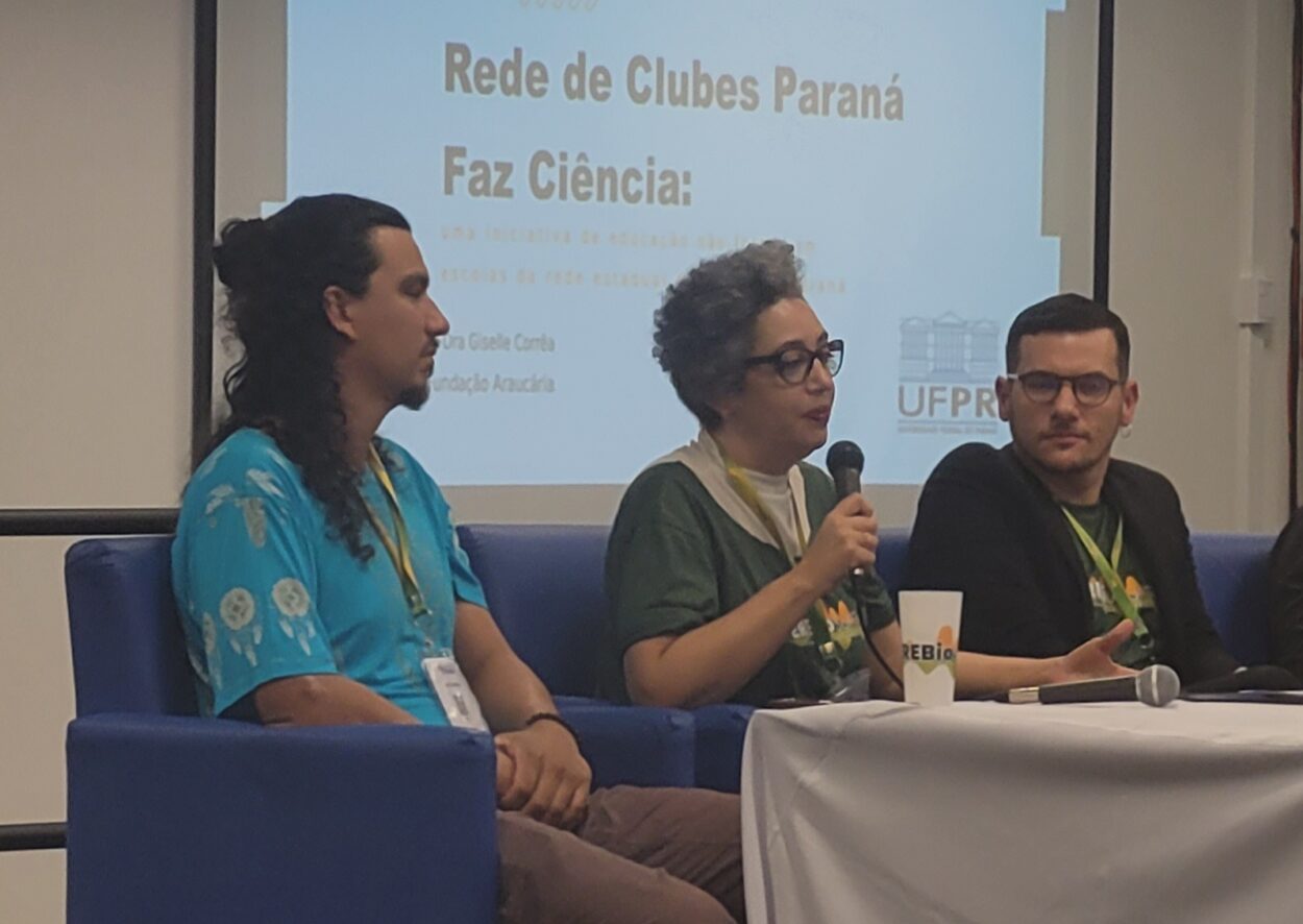 Três pessoas estão sentadas lado a lado em poltronas azuis, participando de uma mesa de conversa. Ao centro, uma mulher de cabelos curtos e grisalhos, usando óculos e camiseta verde da Rede de Clubes Paraná Faz Ciência, segura um microfone enquanto fala. À sua esquerda, um homem de cabelos longos e presos, com camiseta azul e crachá no pescoço, escuta atentamente. À direita, outro homem de barba e óculos, veste blazer preto e camiseta verde, observando a fala da colega. À frente deles, há uma mesa com toalha branca e microfone. No fundo, um telão exibe a apresentação com o título “Rede de Clubes Paraná Faz Ciência” e o logotipo da UFPR.