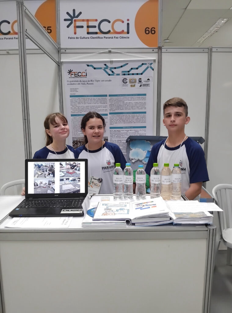 A galeria mostra 3 momentos do Clube de Ciências Aquavida na Feira de Cultura Científica Paraná Faz Ciência (FECCI). O ambiente é coberto e bem iluminado, com estandes e objetos para apresentação da pesquisa desenvolvida pelos clubistas. A última foto mostra o professor Samuel Moraes e os clubistas cujo projeto foi premiado na feira com o 3º lugar na categoria Desenvolvimento de Produtos Junior.