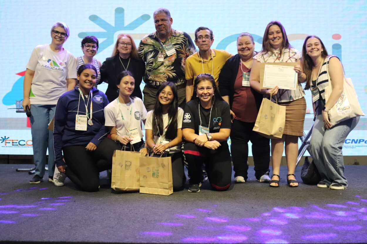 A imagem mostra os integrantes do clube Litterae Ludus durante a entrega do prêmio da categoria a qual se inscreveram para apresentar o trabalho na Fecci. Atrás, há um painel luminoso. Os alunos estão uniformizados e seguram sacolas, troféus e medalhas. Ao lado da equipe, há autoridades, coordenadores e bolsistas pedagógicos. Todos estão em cima de um palco e olham para a frente, posando para a câmera.