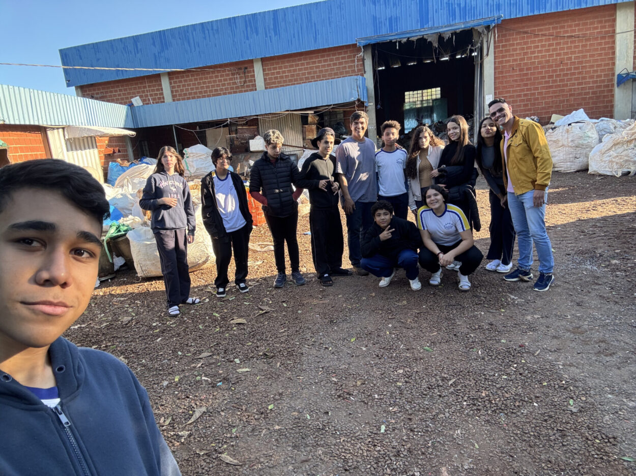 A primeira imagem mostra um grupo de pessoas reunidas em frente a um galpão de tijolos à vista com cobertura azul. Ao fundo, há grandes sacos de materiais, organizados em montes. No centro, está a maior parte do grupo, formado por jovens e um adulto. Duas pessoas estão agachadas na frente, enquanto as demais estão em pé, lado a lado, sorrindo e olhando para a câmera. À esquerda, em primeiro plano, aparece um jovem tirando a foto em modo selfie, capturando todo o grupo. O ambiente é externo, com chão de pedrinhas e iluminação natural, indicando que a foto foi tirada durante o dia. A segunda imagem mostra um grupo de estudantes posando para uma foto ao lado de um ônibus escolar amarelo e branco. A maioria veste uniformes escolares. O grupo parece estar em uma atividade externa. Ao fundo, há um prédio branco com grades e a inscrição verde “CTR”, além de uma torre cilíndrica branca que faz parte da estrutura do local. O céu está limpo e azul, com algumas nuvens espalhadas, indicando que a foto foi tirada em um dia ensolarado. Os estudantes estão sorrindo e interagindo de forma descontraída.
