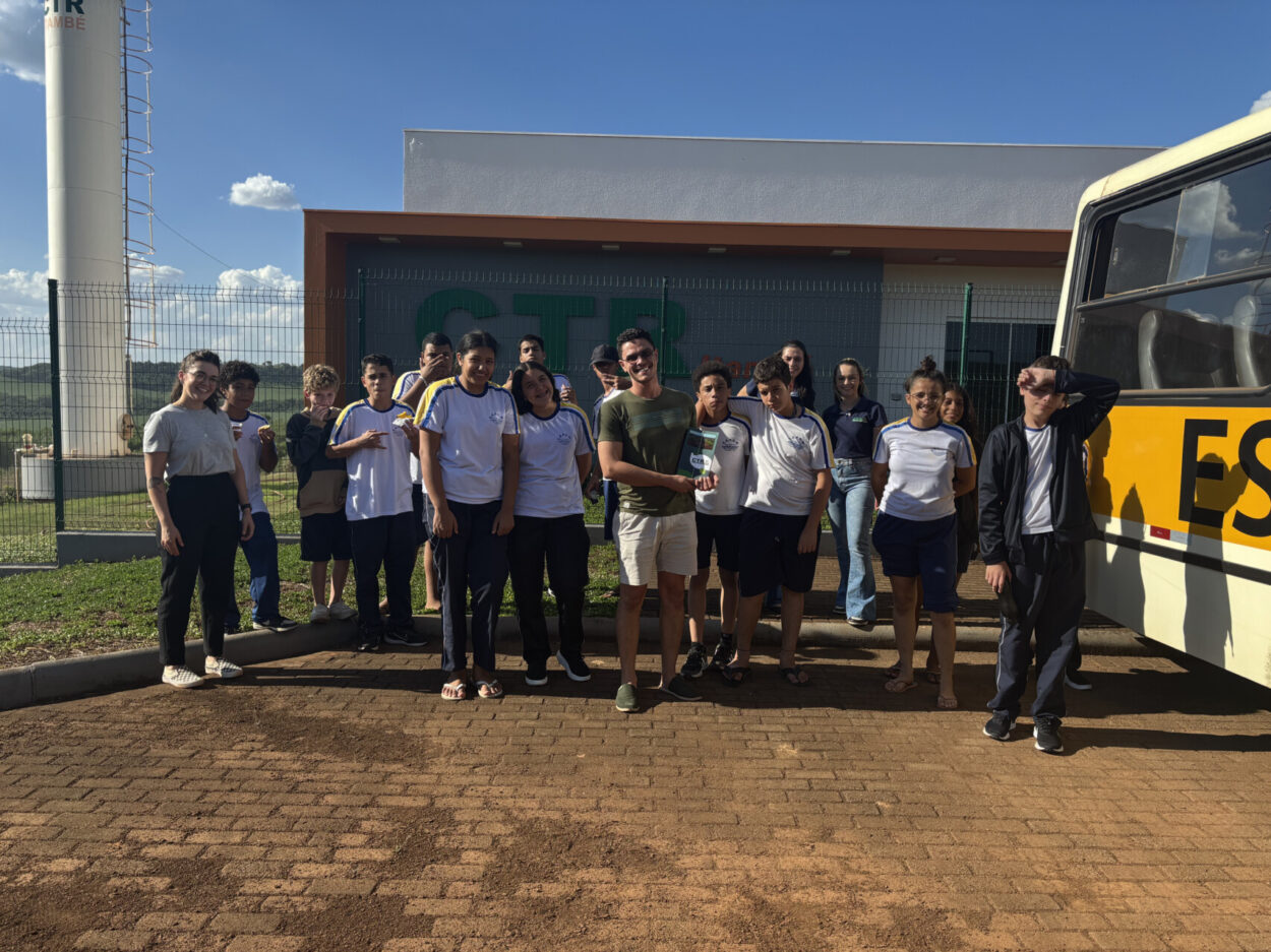 A primeira imagem mostra um grupo de pessoas reunidas em frente a um galpão de tijolos à vista com cobertura azul. Ao fundo, há grandes sacos de materiais, organizados em montes. No centro, está a maior parte do grupo, formado por jovens e um adulto. Duas pessoas estão agachadas na frente, enquanto as demais estão em pé, lado a lado, sorrindo e olhando para a câmera. À esquerda, em primeiro plano, aparece um jovem tirando a foto em modo selfie, capturando todo o grupo. O ambiente é externo, com chão de pedrinhas e iluminação natural, indicando que a foto foi tirada durante o dia. A segunda imagem mostra um grupo de estudantes posando para uma foto ao lado de um ônibus escolar amarelo e branco. A maioria veste uniformes escolares. O grupo parece estar em uma atividade externa. Ao fundo, há um prédio branco com grades e a inscrição verde “CTR”, além de uma torre cilíndrica branca que faz parte da estrutura do local. O céu está limpo e azul, com algumas nuvens espalhadas, indicando que a foto foi tirada em um dia ensolarado. Os estudantes estão sorrindo e interagindo de forma descontraída.