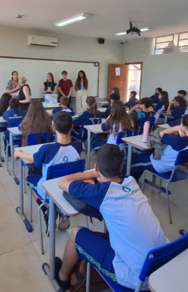 A galeria mostra momentos da oficina “Lixo Eletrônico e Metais Pesados” no Colégio Estadual José de Anchieta, em Dois Vizinhos. Em uma sala de aula com carteiras enfileiradas, lousa e TV para slides, professores demonstram o uso de uma fita indicadora ácido-base em uma mesa com equipamentos.