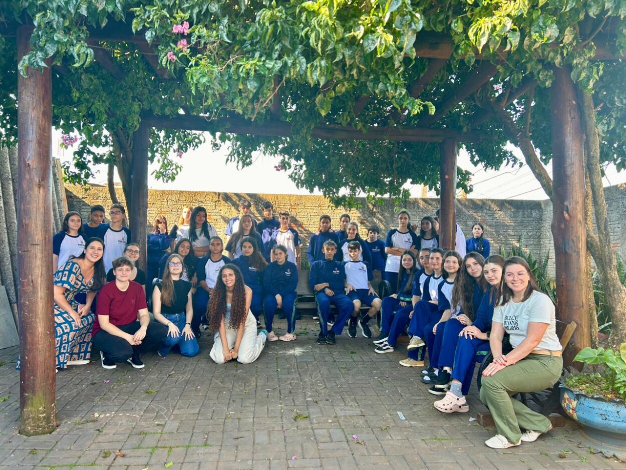 A imagem mostra um grande grupo de estudantes e professoras reunidos em um espaço ao ar livre. Eles posam para a foto sob uma pérgola de madeira coberta por folhas verdes e flores lilases, que criam sombra no ambiente. A maioria usa uniforme azul e branco escolar, e todos sorriem para a câmera. O chão é de tijolos e, ao fundo, há um muro de pedra e vegetação. A cena transmite acolhimento e espírito de grupo.