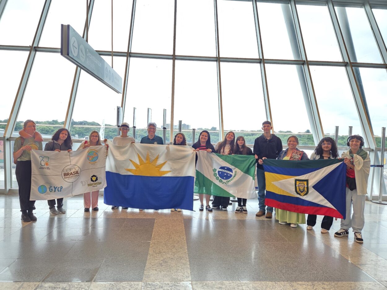 Um grupo de estudantes no saguão de um aeroporto, enfileirados lado a lado, seguram quatro bandeiras no total, uma é do evento COP30 com símbolos da Rede de Clubes Paraná Faz Ciência, do projeto de extensão da Unicentro: Nós Propomos!, do Global Youth Climate Pact (GYCP) - Pacto Mundial de Jovens pelo Clima no Brasil e a logo da Unicentro. Outras bandeiras também são mostradas pelos alunos, como a do Estado do Paraná, da Cidade de Guarapuava e a do Colégio Estadual Padre Chagas, de Guarapuava.