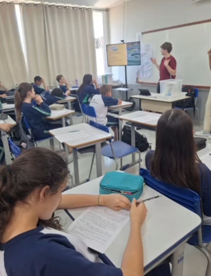 A galeria mostra momentos da oficina “Lixo Eletrônico e Metais Pesados” no Colégio Estadual José de Anchieta, em Dois Vizinhos. Em uma sala de aula com carteiras enfileiradas, lousa e TV para slides, professores demonstram o uso de uma fita indicadora ácido-base em uma mesa com equipamentos.