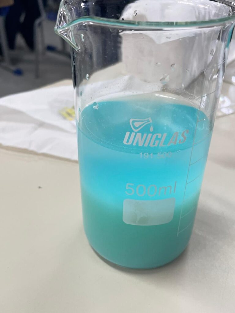 A galeria mostra momentos da oficina “Lixo Eletrônico e Metais Pesados” no Colégio Estadual José de Anchieta, em Dois Vizinhos. Em uma sala de aula com carteiras enfileiradas, lousa e TV para slides, professores demonstram o uso de uma fita indicadora ácido-base em uma mesa com equipamentos.