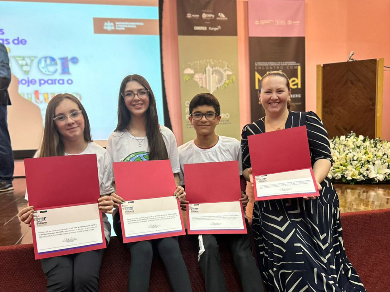 Três estudantes e uma mulher adulta posam sorrindo em um auditório, segurando certificados dentro de pastas vermelhas abertas. Os estudantes usam camisetas brancas e estão sentados em degraus à frente do palco. Atrás deles há um telão com projeções coloridas e banners de eventos científicos. À direita, há um arranjo de flores brancas próximo a um púlpito de madeira. A cena transmite um momento de celebração e premiação.