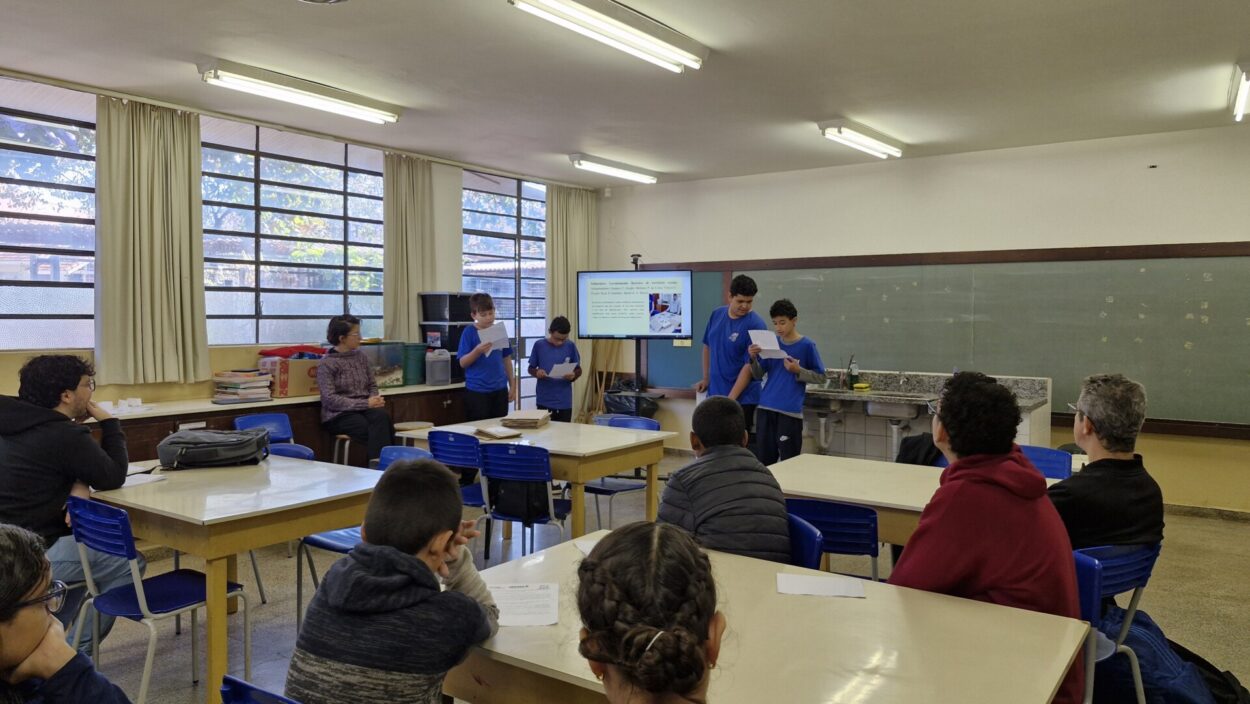 A imagem mostra alguns alunos, com uniformes azuis, apresentando um trabalho durante um seminário. A professora está ao lado, observando enquanto os estudantes explicam a temática estudada. Atrás deles, há uma tela com slides que contém mais informações sobre a investigação. Os alunos estão sendo observados por outros colegas e professores, que prestam atenção durante a explicação.