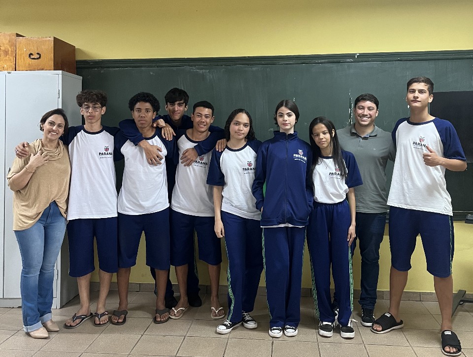 A foto é do Clube de Ciências ‘Green Lab” do Colégio Estadual Almirante Tamandaré de Cruzeiro do Oeste. Na imagem aparece um grupo de nove estudantes e dois adultos dentro de uma sala de aula. Eles estão alinhados lado a lado, formando uma fila, bem próximos uns dos outros. A maioria está usando uniforme escolar azul e branco. Dois adultos — um à esquerda e o outro mais à direita do grupo — também estão presentes. Ao fundo há um quadro verde grande na parede, típico de sala de aula, e a parede é pintada em duas cores: amarelo na parte superior e verde abaixo. O piso é de cerâmica bege, comum em escolas. Estão todos olhando para a câmera e posando para a foto dentro de um ambiente escolar.