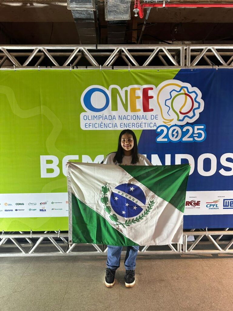 A foto é da aluna Julia Carolina Bunhak Quintino. A imagem mostra a jovem em pé, sorrindo, segurando à sua frente a bandeira do estado do Paraná. Ela está posicionada diante de um grande painel colorido da ONEE – Olimpíada Nacional de Eficiência Energética 2025, que traz o logotipo do evento e a palavra “BEM-VINDOS”. O painel tem cores predominantes em verde, azul e amarelo. A jovem veste calça jeans e tênis escuros. A estrutura metálica acima e abaixo do painel sugere que o local é um espaço de evento. A iluminação destaca tanto a participante quanto a bandeira que ela segura.
