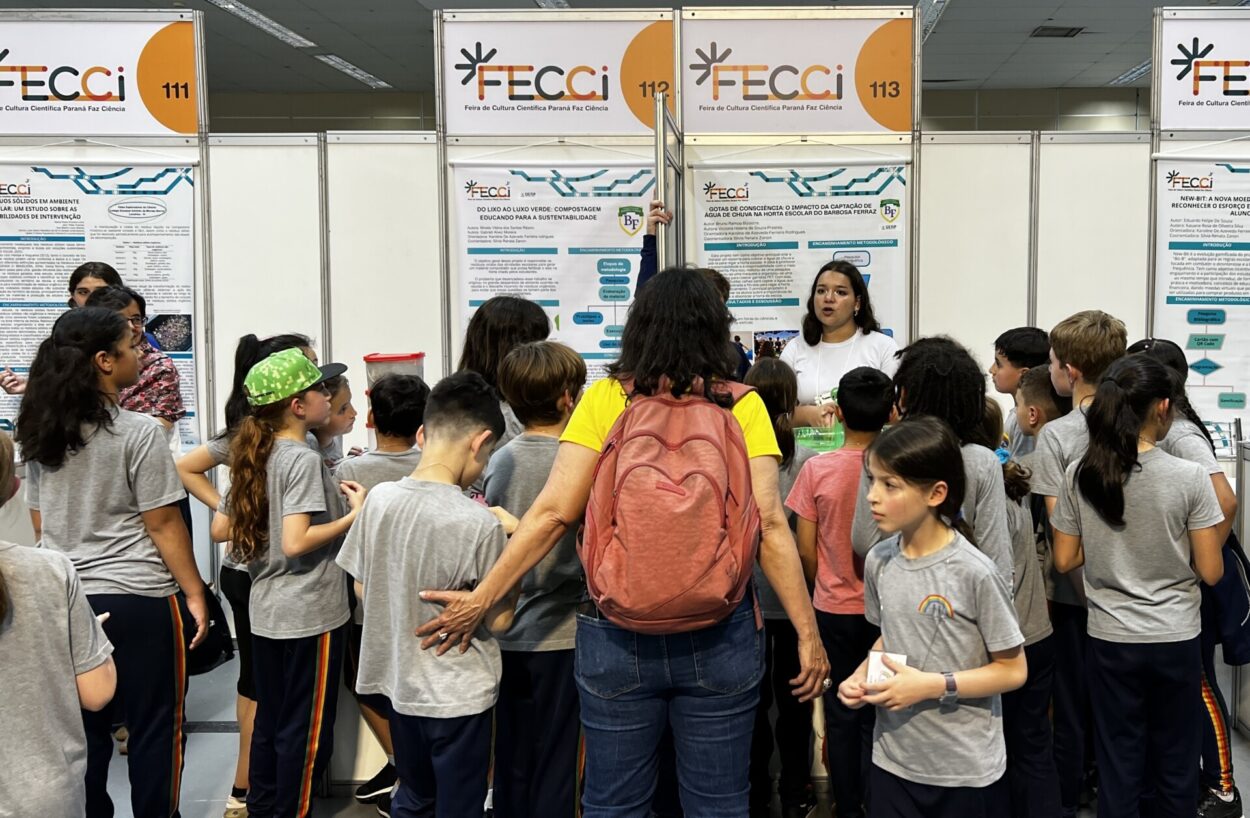 Um grupo de crianças, acompanhadas por uma adulta, observa atentamente a apresentação de uma menina em um estande de feira científica. O espaço tem painéis com cartazes explicativos, identificados como parte da FECCI, e o ambiente é organizado como uma exposição educativa, com várias pessoas reunidas.