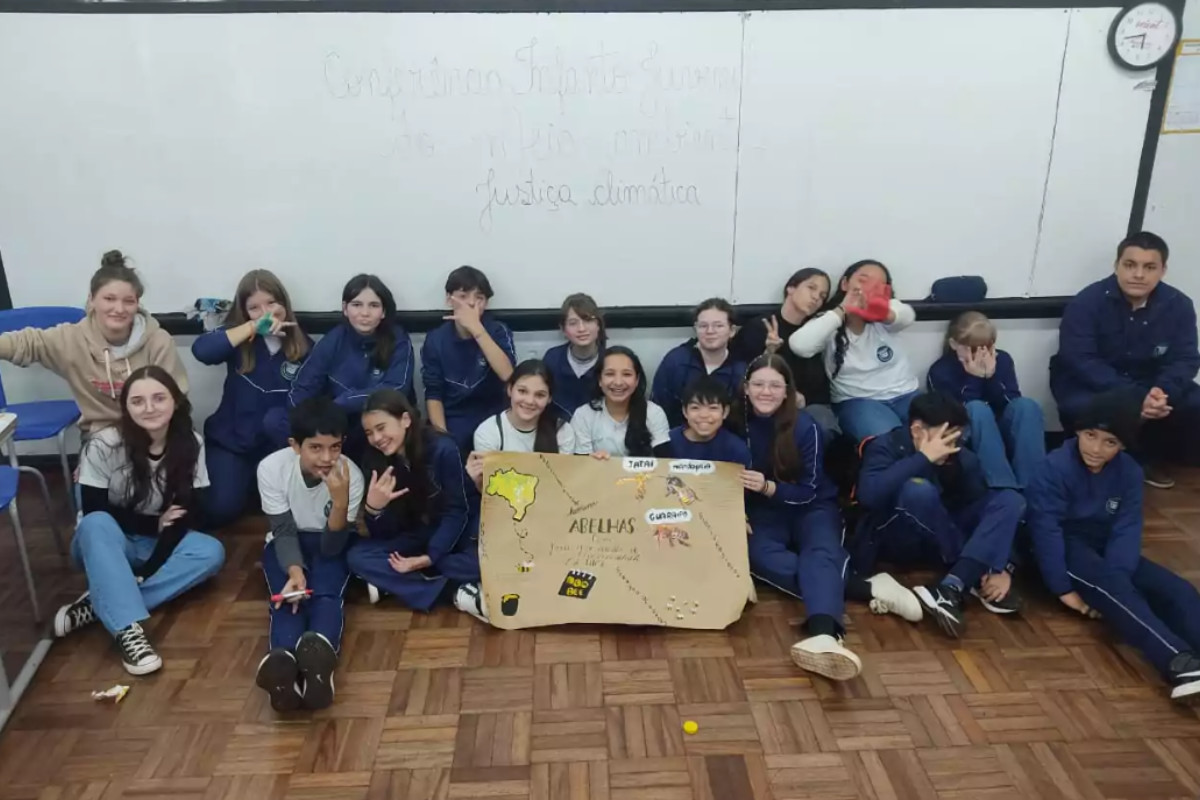 A imagem mostra um grupo de estudantes reunido dentro de uma sala de aula. Eles estão sentados no chão de madeira em frente a um quadro branco, onde está escrito em letras cursivas algo como “Conferência Infanto Juvenil pelo Meio Ambiente – Justiça Climática”. No centro do grupo, algumas crianças seguram um cartaz feito em papel pardo. Nele aparece o desenho do mapa do Brasil, com o título “Abelhas” e algumas palavras como “Jataí”, “Mirimguaçu” e “Guarapu”, que são nomes de espécies de abelhas nativas.