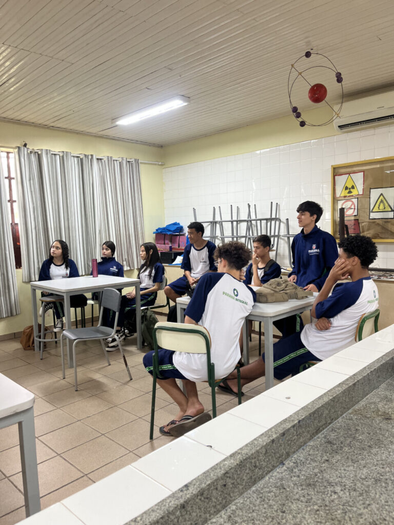 Na imagem aparece um grupo de estudantes dentro de uma sala de aula, organizados em mesas que estão posicionadas próximas umas das outras, formando pequenos agrupamentos. Eles parecem estar prestando atenção em alguém que está fora do quadro da foto. A maioria está com uniforme escolar azul e branco. Eles estão sentados de forma descontraída, mas atentos, demonstrando concentração e interesse. O ambiente é um laboratório escolar: à direita é possível ver cartazes de sinalização de risco, como símbolos de radiação, perigo e proibição. Ao fundo há cadeiras empilhadas sobre uma bancada e materiais escolares. A parede dos fundos é revestida com azulejos brancos e o restante da sala é pintado na cor amarelo claro. O piso é de cerâmica bege.