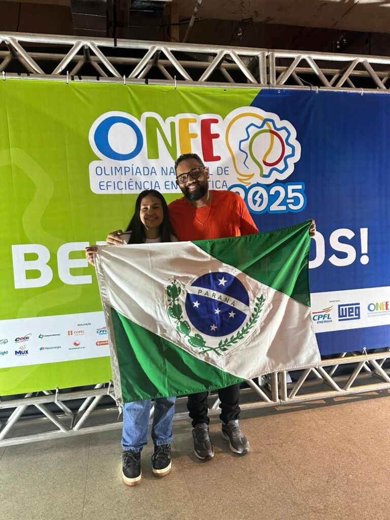 A imagem mostra duas pessoas posando juntas em frente a um grande painel da ONEE – Olimpíada Nacional de Eficiência Energética 2025. À esquerda, está uma jovem sorrindo, e ao lado dela, um homem que apoia o braço sobre o ombro dela, também sorrindo. Ambos seguram, à frente do corpo, a bandeira do estado do Paraná, estendida horizontalmente. O fundo é composto por um painel colorido em verde e azul, com o logotipo da ONEE e a palavra “BEM-VINDOS”. A estrutura metálica superior faz parte do cenário do evento.