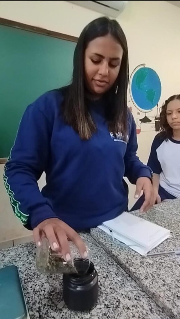 A imagem mostra uma jovem em uma sala de aula realizando uma atividade prática. Ela está em pé ao lado de uma bancada de granito e despeja o conteúdo de um pequeno pote de vidro dentro de um frasco escuro. A estudante veste uma blusa de uniforme escolar azul. Sobre a bancada, há também um caderno ou bloco de anotações e uma caneta. Ao fundo, vê-se parte de um quadro verde e, na parede, um mapa-múndi. Outra estudante aparece à direita, observando a atividade.