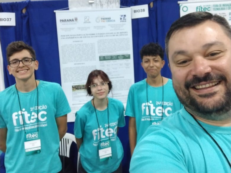 A imagem mostra uma equipe de quatro pessoas, três alunos e um professor. Todos vestem blusas azul turquesa, próprias do evento. A equipe é do clube Unidade Polo e está posando para a foto, sorrindo, tirando uma selfie. Atrás, há um banner com o projeto apresentado pelo grupo.