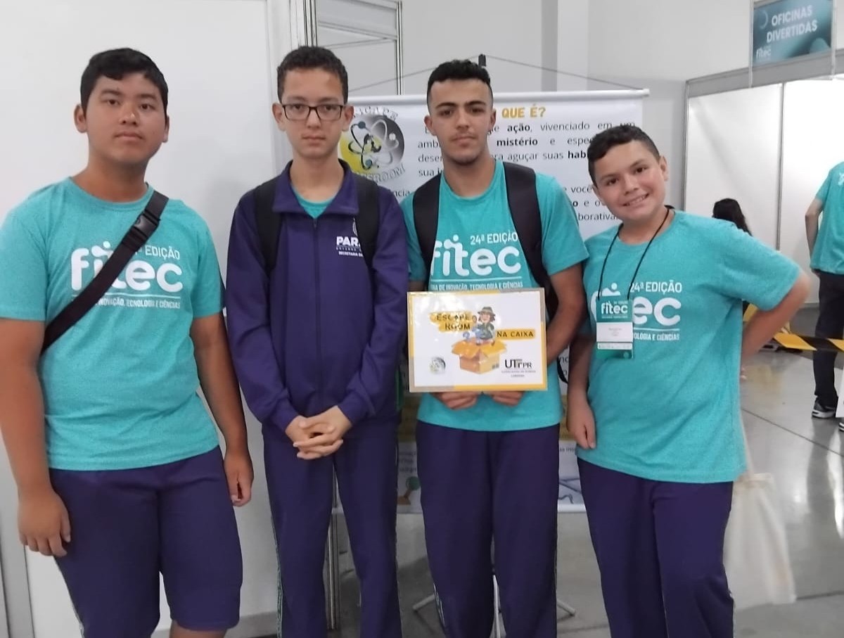 A imagem mostra um grupo de quatro estudantes, pertencentes ao clube Eco Sapiens. Os alunos posam para a foto, vestidos com a blusa do evento, azul turquesa. Um dos estudantes segura um certificado, recebido pelo segundo melhor tempo no escape room, uma das atividades ofertadas na Feira.