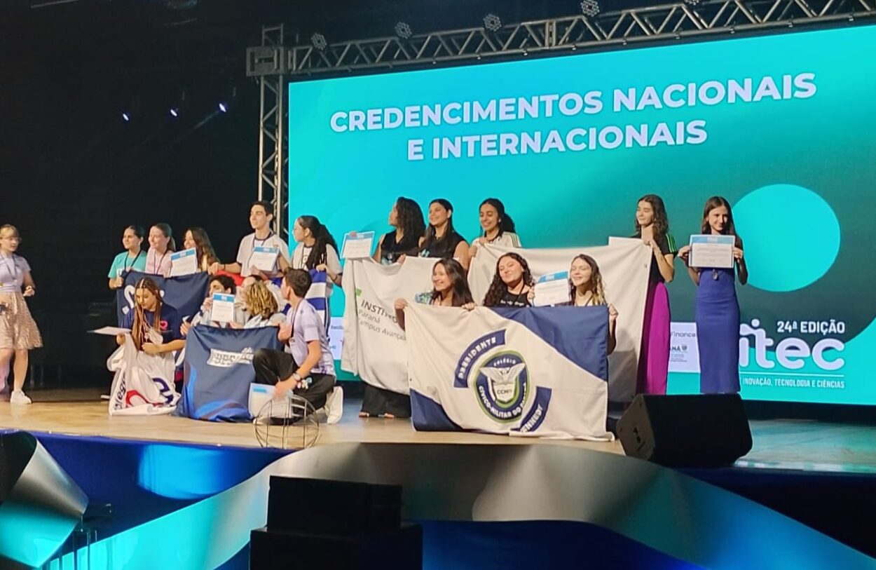 A imagem mostra um momento importante, em uma premiação. Atrás, há um painel luminoso indicando que é o momento de entregar o credenciamento para outra feira. No palco, há várias estudantes segurando bandeiras das escolas as quais fazem parte. No meio, há o grupo do clube Discovery Kennedy’s, que recebeu dois certificados pelo projeto apresentado no evento.