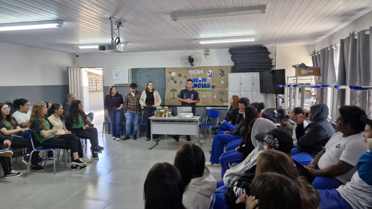 A imagem mostra uma sala de aula ampla e bem iluminada, com estudantes sentados, atentos a uma apresentação. Na frente, a equipe NAPI/ UEL está em pé atrás de uma mesa, falando e mostrando frascos com líquidos e materiais científicos. Na parede atrás deles, há um cartaz grande com o título “Clube de Ciências” e desenhos relacionados à natureza. O público parece bastante interessado e o ambiente transmite uma sensação de engajamento e troca de conhecimento.