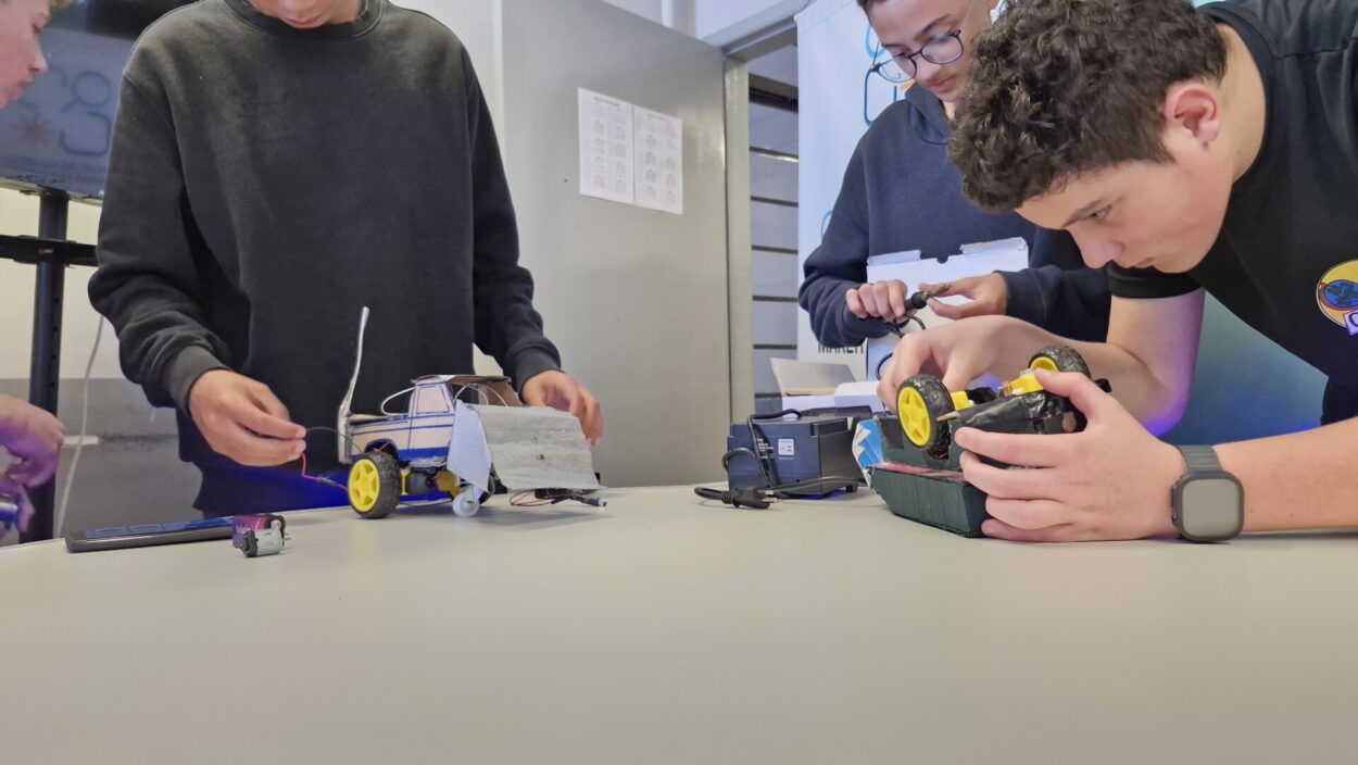 A imagem mostra alunos reunidos em uma mesa de trabalho, concentrados em montar robôs feitos com materiais simples, como papelão, fios e rodas plásticas amarelas. Dois deles observam e ajustam peças elétricas nos veículos, enquanto outro parece testar uma parte com um fio conectado. O ambiente é uma sala de aula, com paredes cinza e um armário metálico ao fundo.