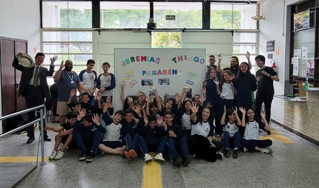 Fotografia em ambiente interno de uma escola. No centro da imagem, há um grande painel branco decorado com letras coloridas formando a frase “Jeremias Thiago Parabéns”, acompanhado de pequenos balões de fala com mensagens e fotos das atividades do clube. Em frente ao painel, sentados no chão, está um grupo numeroso de estudantes, a maioria usando uniforme azul e branco. Atrás deles, outros estudantes estão em pé ao lado do painel. À esquerda, aparece um homem de chapéu claro, camisa social e gravata, e outro homem de camiseta azul. À direita, há mais professores e estudantes em pé. O grupo posa para a foto em clima de celebração, sugerindo um momento de homenagem ou reconhecimento ao trabalho realizado pelo clube na escola.
