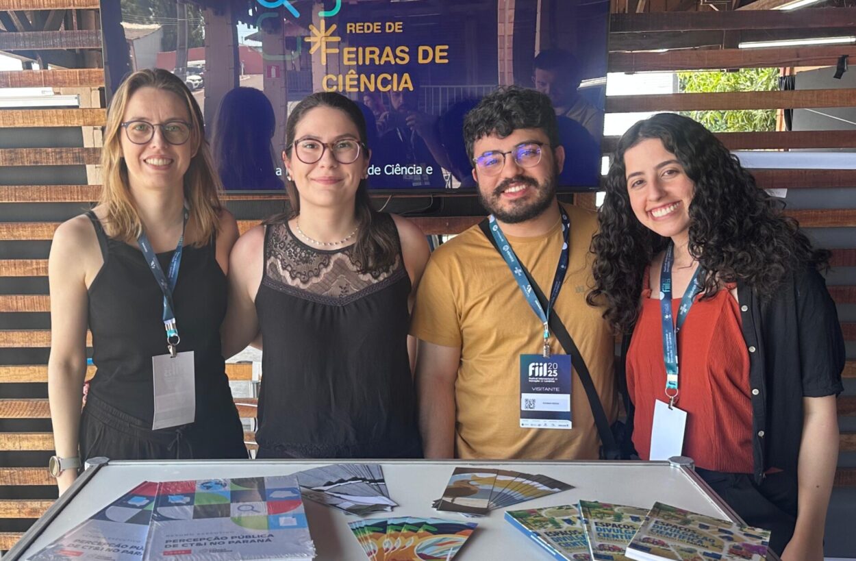 A imagem mostra um grupo de quatro pessoas, todos membros da equipe do NAPI PRFC que compareceu ao evento. Atrás, há uma televisão com um vídeo institucional. No balcão na frente, há livros, marca-páginas e a pesquisa de percepção pública desenvolvida pelo NAPI. Todos sorriem e posam para a foto.