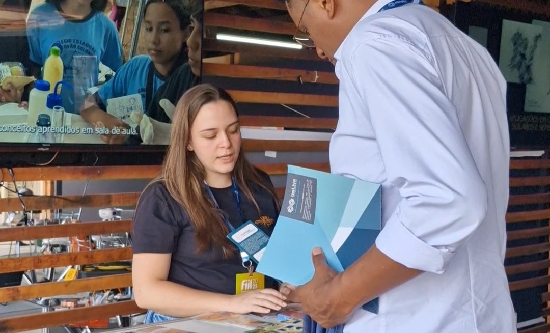 A imagem mostra uma das integrantes da equipe do NAPI PRFC apresentando o projeto para um homem alto. Atrás dela, há uma televisão com um vídeo institucional. No balcão em sua frente, há livros, marca-páginas e a pesquisa de percepção pública desenvolvida pelo NAPI. O homem segura um livro também, sobre espaços de divulgação científica.
