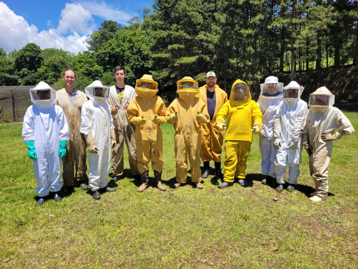 Dez pessoas em pé posam para a foto, elas estão trajando macacões de segurança apícola, de forma que se cobre todo o corpo dos pés a cabeça, com visor característico do equipamento. O espaço ao redor é verdejante, com árvores ao fundo e céu azul.