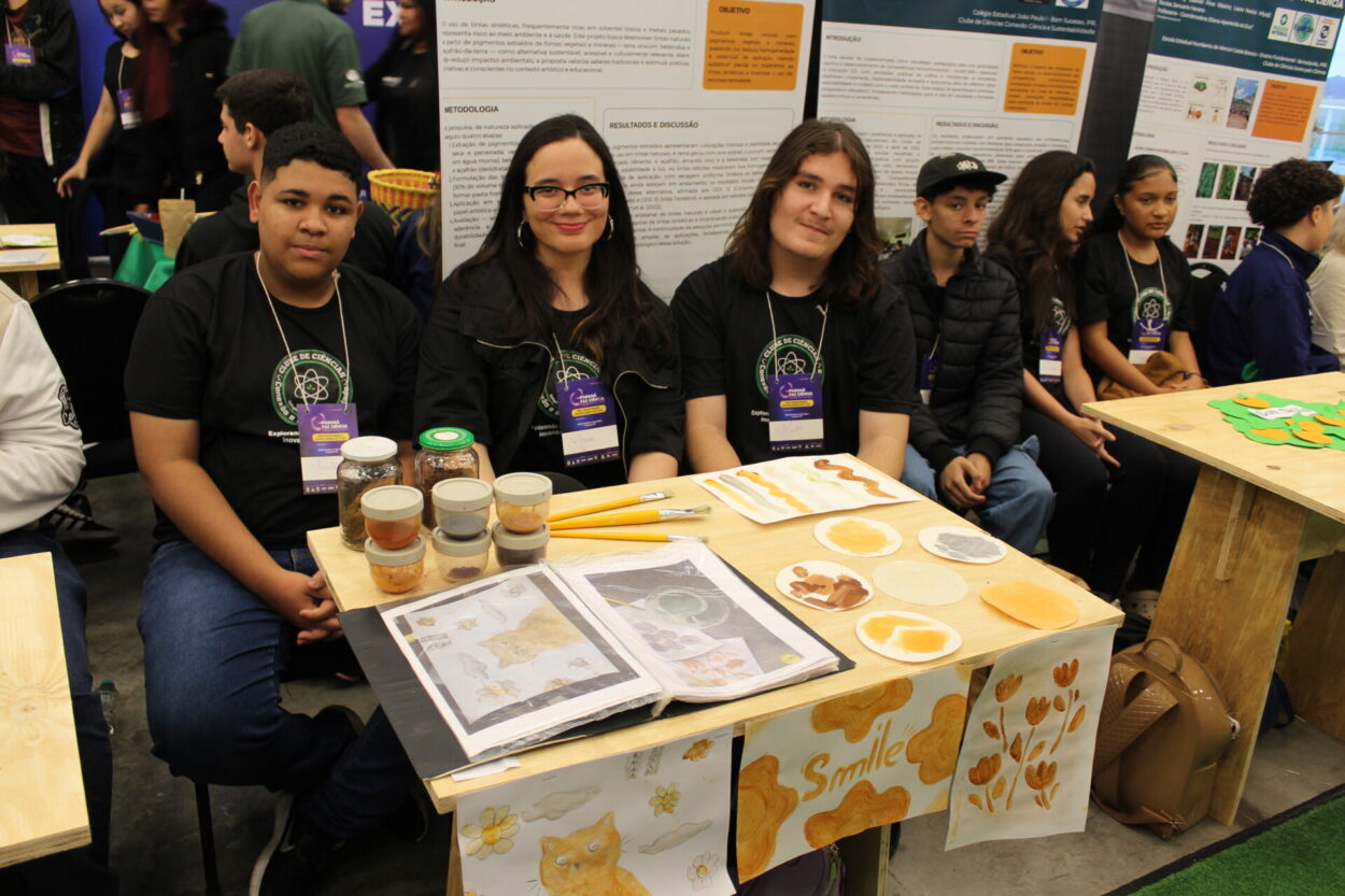 Três estudantes estão sentados atrás de uma mesa de exposição em um evento científico. Eles vestem camisetas pretas com o logotipo de um clube de ciências e crachás roxos no pescoço. À frente deles, a mesa exibe potes de vidro com pigmentos naturais, pequenas amostras de tintas artesanais em tons terrosos, pincéis e folhas com pinturas de gatos, flores e círculos coloridos. A mesa também tem cartazes decorados com desenhos em tons de marrom e laranja, incluindo a palavra “Smile”. Atrás do grupo, há painéis de pesquisa e outros estudantes sentados, indicando que o evento está movimentado.