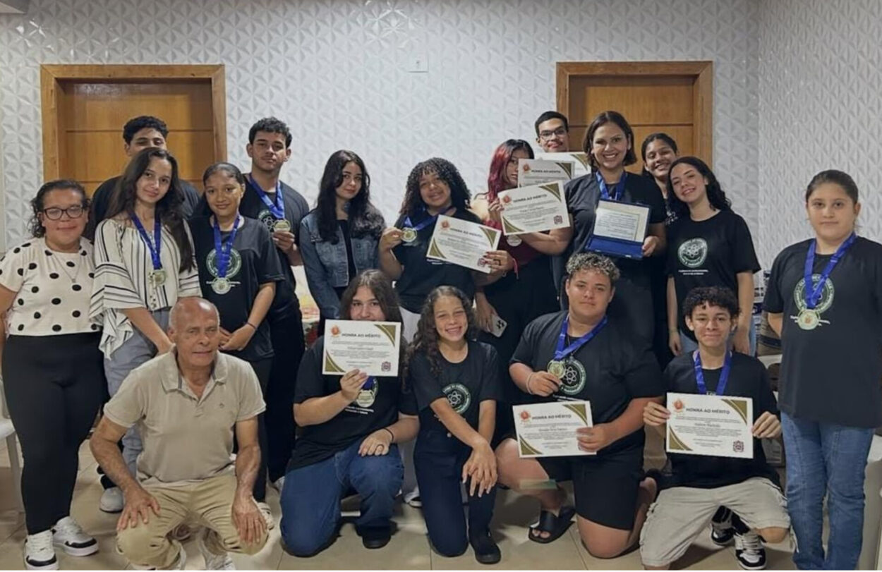 Um grupo de pessoas, entre adolescentes e alguns adultos, posa em uma sala com paredes claras e textura geométrica. A maioria usa camisetas pretas com o logotipo de um clube de ciências e está com medalhas azuis no pescoço. Vários deles seguram certificados, sorrindo para a foto. Na frente, algumas pessoas estão agachadas ou sentadas, também exibindo certificados. O clima é de comemoração e conquista coletiva.