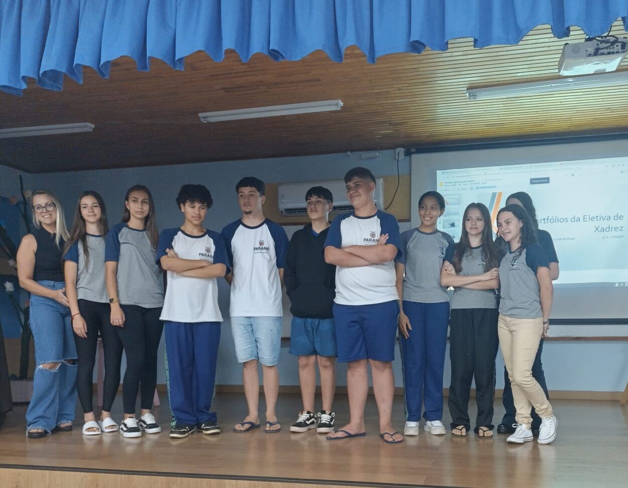 Um grupo de pessoas, composto por uma professora e estudantes, está posicionado em um palco de madeira dentro de uma sala escolar. Eles estão alinhados lado a lado, alguns com os braços cruzados e outros com as mãos ao lado do corpo. A maioria usa uniformes escolares em tons de azul, branco e cinza. Ao fundo, há uma cortina azul e, no lado direito, um projetor que exibe o portfólios de projetos dos alunos. Todos com expressões tranquilas e sorridentes, reunidos para uma apresentação escolar.