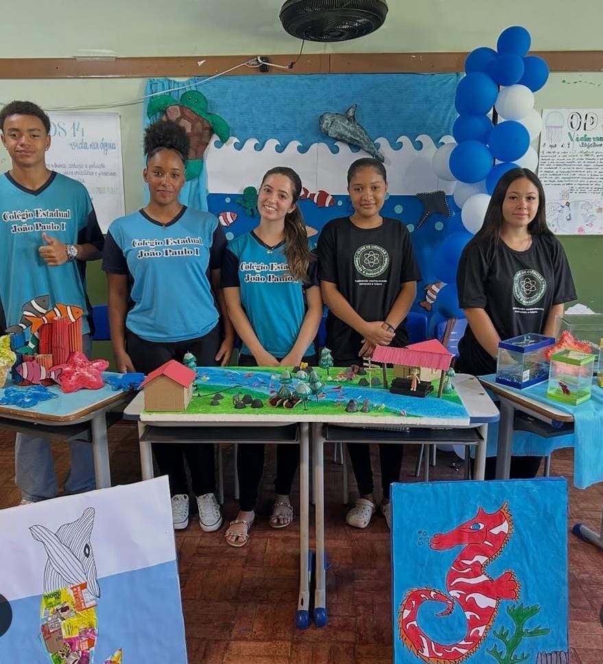 Cinco estudantes estão em pé atrás de uma mesa em uma sala de aula decorada com o tema “vida marinha”. Três deles vestem camisetas azuis com o nome do Colégio Estadual João Paulo I, e dois vestem camisetas pretas com o logotipo de um clube de ciências. Sobre as mesas à frente, há maquetes coloridas representando ambientes aquáticos, casas, árvores e animais marinhos. Ao fundo, um painel azul mostra desenhos de peixes, ondas, uma tartaruga e um golfinho, além de balões azuis formando um arco. Nas laterais, dois cartazes artísticos exibem um desenho de cavalo-marinho e a silhueta de um peixe preenchida com recortes coloridos. O grupo sorri levemente, posando para a foto.