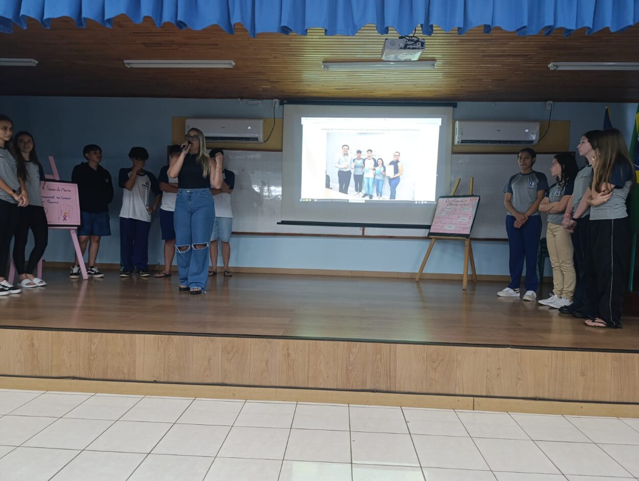 A galeria registra momentos da Culminância do Colégio Estadual Guilherme de Almeida, em Santa Izabel do Oeste (PR). Em um auditório com carteiras enfileiradas, palco, cortinas e projetor, professores e clubistas apresentam os projetos desenvolvidos ao longo do ano de 2025. Há, ainda, imagens de um panfleto intitulado 'Agrotóxicos e o Câncer de Mama' e outros momentos marcantes do clube.