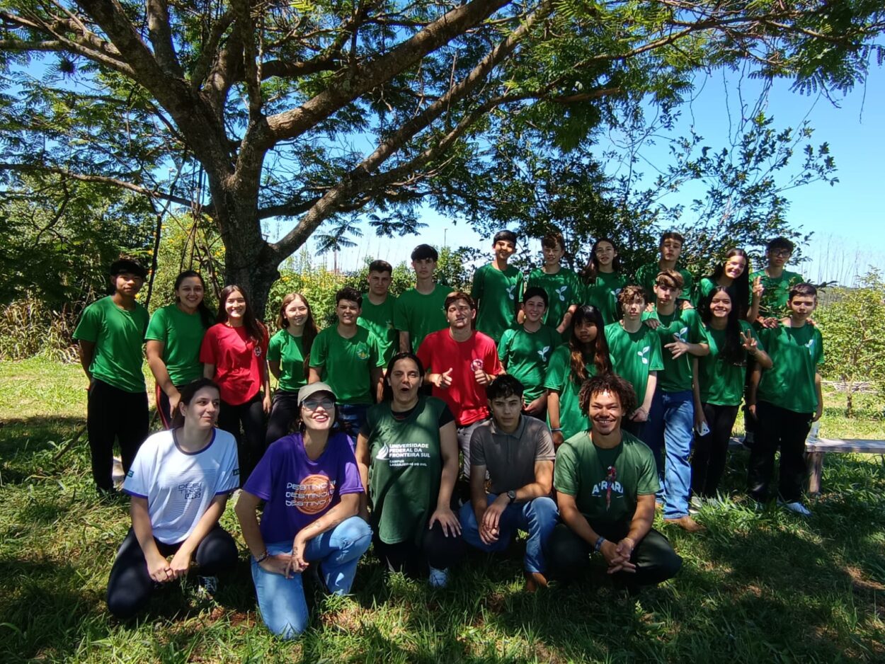 Foto dos clubistas do Meu Mundo Melhor em visita a UFFS e após oficina sobre pomares e hortas. Na imagem, 20 alunos posam para a foto de pé e 5 integrantes do projeto de extensão da Universidade estão abaixados a sua frente, também olhando para a câmera. A maioria demonstra alegria, os estudantes estão todos uniformizados. O ambiente é sombreado por uma grande árvore ao fundo, o solo é coberto por grama e no horizonte um céu azul e mais plantas altas.