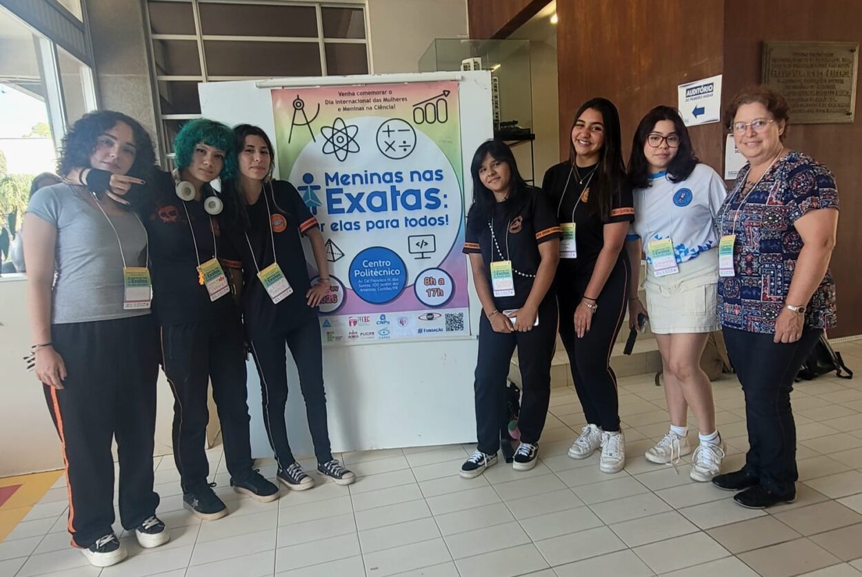 Sete meninas adolescentes e uma professora posam em pé ao lado de um banner do evento “Meninas nas Exatas”, no Campus Politécnico da UFPR. Elas usam crachás e camisetas do clube, em ambiente interno da universidade.