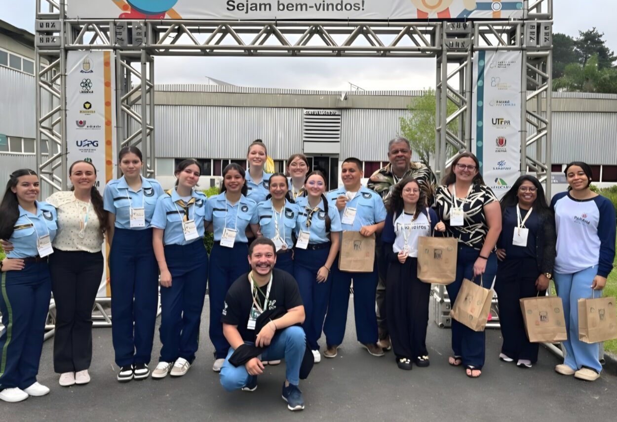 A imagem mostra parte da equipe da UNILA e dos clubes de ciências articulados pela instituição. Na foto, o grupo está posicionado em frente ao painel de boas-vindas da feira FECCI. Os alunos estão uniformizados, ao lado do professor Ronaldo, do bolsista Lucas e das professoras coordenadoras dos clubes. Há três clubes na imagem.