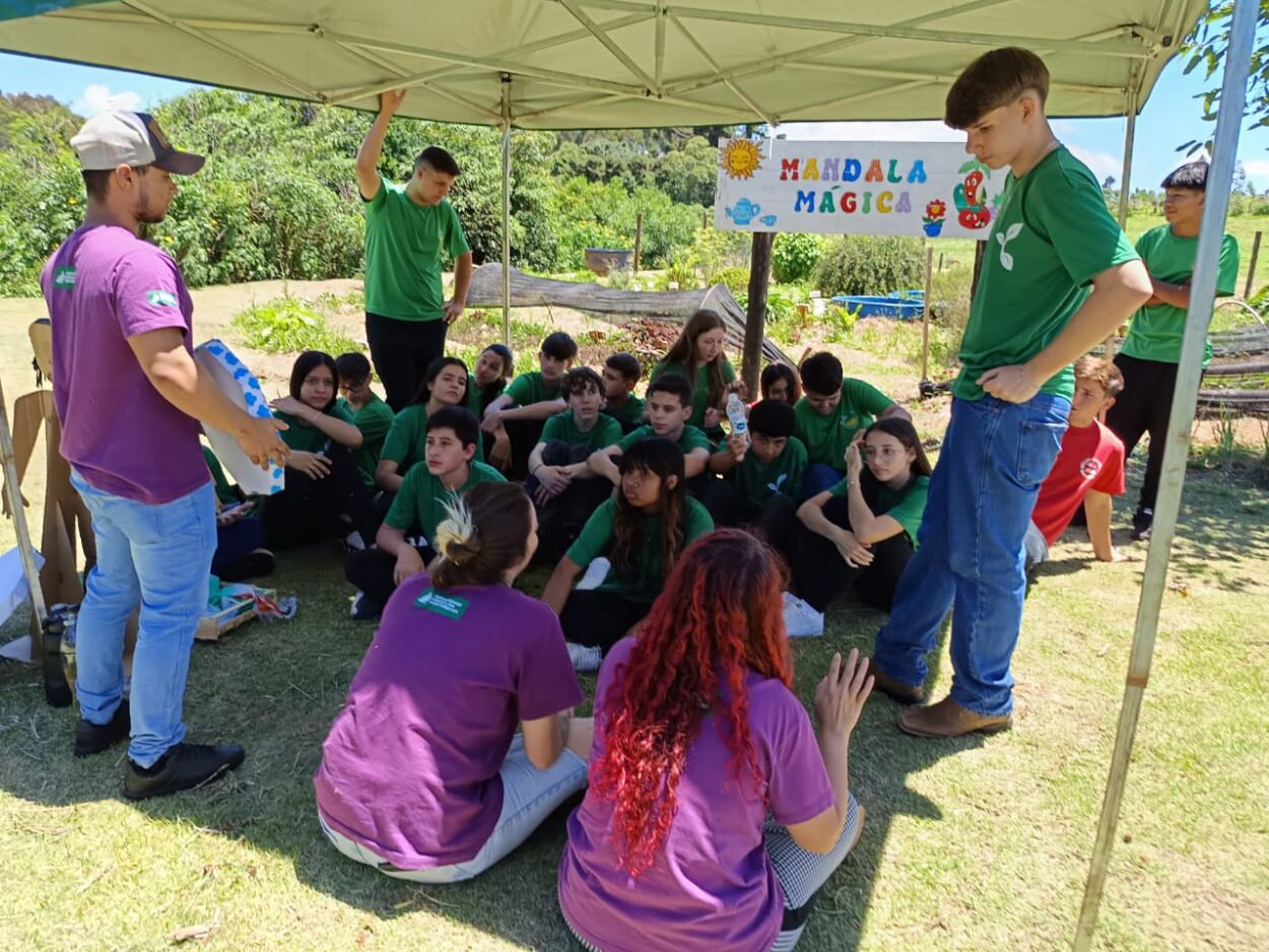 Sobre a sombra de uma tenda, em sua maioria sentados, os clubistas observam os três oficineiros - de costas para a câmera - em momento expositivo da oficina. Ao fundo da foto, um espaço de mata fechada e, mais próximo ao grupo, uma horta, e a placa colorida “mandala mágica”. O ambiente exala curiosidade por parte dos estudantes.