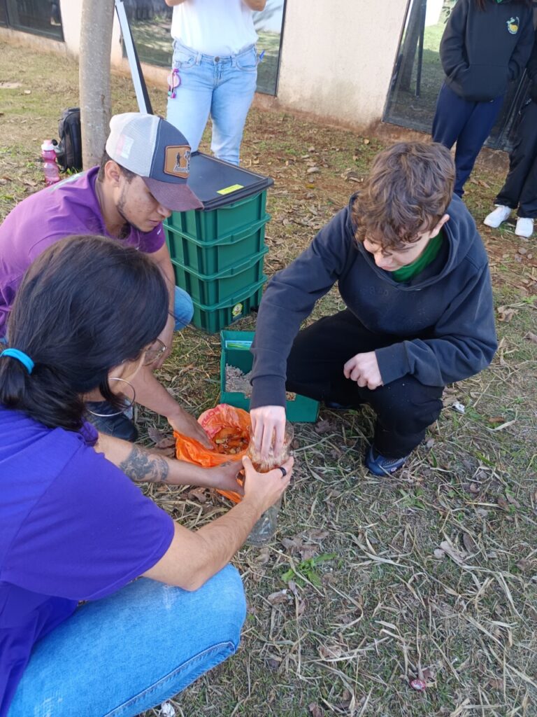 Três pessoas estão agachadas manipulando materiais orgânicos sobre o chão, entre elas um clubista. Uma delas segura um recipiente transparente enquanto outra coloca materiais dentro dele, e a terceira auxilia no processo. Ao fundo, há um conjunto de caixas verdes empilhadas e outras pessoas em pé, parcialmente visíveis. A cena ocorre em um espaço externo com grama seca e folhas espalhadas.