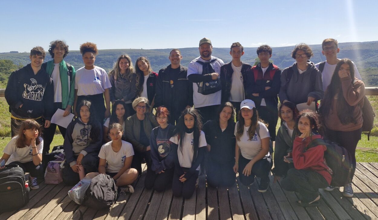 A imagem mostra um registro coletivo ao ar livre. Os estudantes e dois adultos estão organizados em duas fileiras sobre um deck de madeira. Atrás deles aparece uma vista extensa de vales e morros cobertos por vegetação. A iluminação indica um dia claro, com sol forte. Alguns alunos seguram folhas de papel e vários têm mochilas apoiadas no chão, sugerindo uma visita educativa ou excursão. O grupo está próximo, olhando para a câmera, com expressões tranquilas e satisfeitas.
