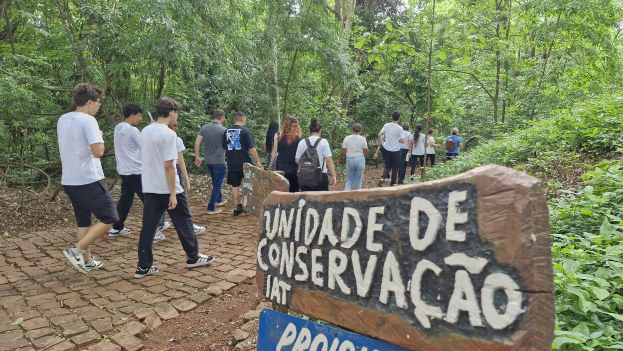 A imagem mostra um grupo de estudantes caminhando por uma trilha em meio a uma área de mata densa e verde. Eles seguem por um caminho de pedras e terra, vistos principalmente de costas, como se estivessem iniciando ou continuando uma caminhada guiada. No primeiro plano há uma placa de madeira com a inscrição “Unidade de Conservação”, indicando que o local é uma área protegida. As árvores são altas e a vegetação é abundante, criando uma atmosfera natural e educativa, típica de atividades de campo voltadas ao aprendizado ambiental.